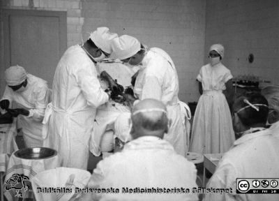 Operation på 1930-talet, troligen i Lund
Omonterat fotografi. Påskrift: "Från Elof Munck af Rosensköld under studietiden på 1930-talet. Aseptik."
Notera att besökare / åskådare visserligen har vita överdragskläder och munskydd, men de har ingen mössa. Assisterande icke steril personal har däremot mössa. 
Nyckelord: Kirurgi;Operation;Demonstration;Aseptik;1930-talet;Foto;Omonterat;Kapsel 08