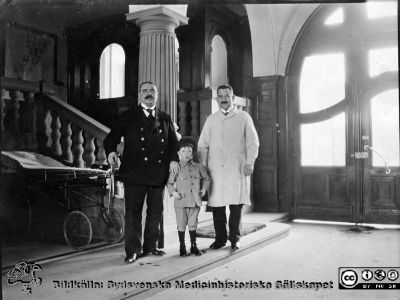 Portvakten Nils Nilsson på Malmö Allmänna Sjukhus, i portgången
Portvakten Nils Nilsson (1874 - 1944) i ljus rock på sin arbetsplats i portgången vid huvudentrén på Malmö Allmänna Sjukhus tillsammans med en uniformerad tjänsteman och en en liten pojke.  Foto ungefär 1920. Bildkälla: Bengt Carlsson, Lotsgatan 208, Limhamn.
Nyckelord: MAS;UMAS;Malm;Allmämma;Sjukhus;Portvakt;Administration
