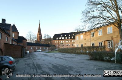 En bakgård år 2015 som en gång var framsidan på Lasarettet i Lund
Livets Museum, fasader mot norr och öster. Mark- och Parktjänst har lokalerna i bottenpanet. Museets entré skymtar  längst bort vid Rehabhuset. 
Till höger en anskrämlig barack för telefonväxlel mm. I bakgrunden Allhelgonakyrkan. 
Denna lasarettets fula bakgård var en gång dess stolta framsida, och kontrasterar markant mot den fina parken bredvid framför UB. Denna ägs och sköts av Lunds Universitet. Där är sikten fri och pryds av moderna statyer och avgränsas av vackra fasader. 
Region Skåne markerar här med eftertryck sin avsaknad av intresse för skönhetsvärden.
Nyckelord: Lasarett;Lund;Universitet;Universitetssjukhus;USiL;Museum;Mark och Park;Telefonväxel;Kyrka;Rehabilitering