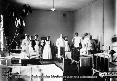 Professor Seved Ribbing går rond 1910 på medicinkliniken i Lund. 
Få¥n samma negativ som SMHS6243_000_01.tif. Publicerad på sid. 199 i Flaum 1968. Bildtext där: "Nu finns elektrisk belysning och värmeledning. Kaminer och kakelugnar togs bort hÃ¤r Ã¥r 1904. Avdelningssköterskan Laura Pedersen utexaminades med den första SSSH-kursen år 1905. Märk de svarta sängtavlorna, påskrivna med krita".  
Nyckelord: USiL;Lund;Universitet;Sjukhus;Medicin;Rond;Klinik