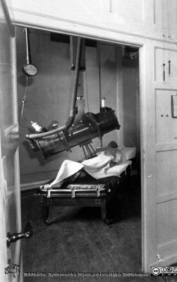 Böntgenbehandling i Lund efter 1931.
På röntgeninstitutet i Lund installerades 1928 - 1931 bildens helkapslade, takhängda röntgenrör och man skärmade av behandlingsrummets väggar med blyplåt för att skydda personalen, som fick sitta utanför och observera patienten genom ett skyddande blyglasfönster. Den bullrande elektriska utrustningen sattes i ett eget utrymme.
Nyckelord: Lasarett;Lund;Sjukhus;Radiologi;Röntgen;Röntgenterapi;USiL;Universitet