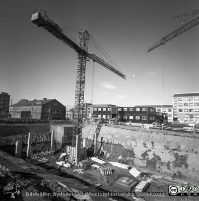 Cetralblocket i Lund byggs 1965
Pärm "Div. tagningar, 1960 och t.v.". Mars, 1965.	Centralblocket på Lasarettet i Lund har just börjat byggas. Längt till vänster i bakgrunden barnkliniken, sedan centraltandpolikliniken (närmare), administrationsbyggnaden (mest skymd), byggbaracker och AB-blocket. Från negativ.
Nyckelord: Lund;Lasarett;Universitet;Universitetssjukhus;Centralblocket;USiL;Bygge;Grundläggning