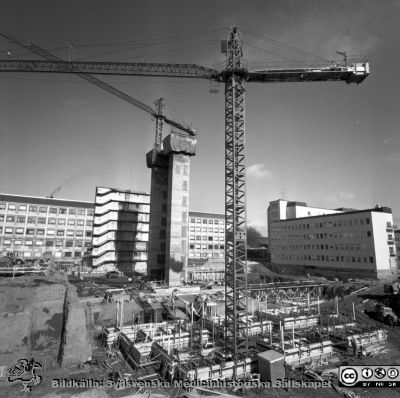 Cetralblocket i Lund byggs 1965
Pärm "Div. tagningar, 1960 och t.v.". Mars, 1965. Centralblocket på Lasarettet i Lund har just börjat byggas. I bakgrunden det tidigare byggda AB-blocket, som fått söderväggen på sitt trapphus avskalad. Strax hitom ett glidformsgjutet betongtorn för det norra hissbatteriet. Till höger E-blockets redan byggda första flyglar. I förgrunden läggs grunden för det södra hissbatteriets torn. Från negativ.
Nyckelord: Lund;Lasarett;Universitet;Universitetssjukhus;Centralblocket;USiL;Bygge;Grundläggning;Hissbatteri;Torn