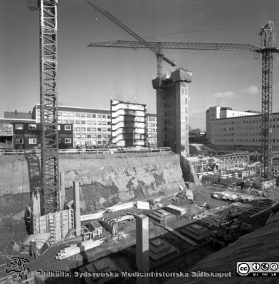 Cetralblocket i Lund byggs 1965
Pärm "Div. tagningar, 1960 och t.v.". Mars, 1965. Centralblocket på Lasarettet i Lund har just börjat byggas. Byggbaracker i bildens vänstra kant. I bakgrunden det tidigare byggda AB-blocket, som fått söderväggen på sitt trapphus avskalad. Strax hitom ett glidformsgjutet betongtorn för det norra hissbatteriet. Till höger E-blockets redan byggda första flyglar. I förgrunden läggs grunden för det södra hissbatteriets torn och D-flyglarna. Från negativ.
Nyckelord: Lund;Lasarett;Universitet;Universitetssjukhus;Centralblocket;USiL;Bygge;Grundläggning;Hissbatteri;Torn