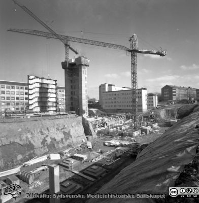 Cetralblocket i Lund byggs 1965
Pärm "Div. tagningar 1960 och t.v.". Mars, 1965. Centralblocket på Lasarettet i Lund har just börjat byggas. Till vänster i bakgrunden det tidigare byggda AB-blocket, som fått söderväggen på sitt trapphus avskalad. Strax hitom ett glidformsgjutet betongtorn för det norra hissbatteriet. Till höger E-blockets redan byggda första flyglar. I bakgrunden till höger om dem psykiatriska kliniken. I förgrunden läggs grunden för det södra hissbatteriets torn och D-flyglarna. Från negativ.
Nyckelord: Lund;Lasarett;Universitet;Universitetssjukhus;Centralblocket;USiL;Bygge;Grundläggning;Hissbatteri;Torn