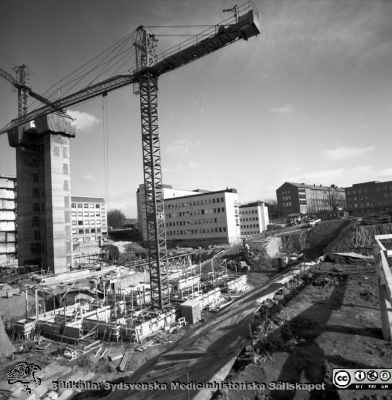 Cetralblocket i Lund byggs 1965
Pärm "Div. tagningar 1960 och t.v.". Mars, 1965. Centralblocket på Lasarettet i Lund har just börjat byggas. Till vänster i bakgrunden dskymtar et tidigare byggda AB-blocket, som fått söderväggen på sitt trapphus avskalad. Strax hitom ett glidformsgjutet betongtorn för det norra hissbatteriet. Till höger E-blockets redan byggda första flyglar. I bakgrunden till höger om dem psykiatriska och barnpsykiatdriska klinikerna. I förgrunden läggs grunden för det södra hissbatteriets torn och D-flyglarna. Från negativ.
Nyckelord: Lund;Lasarett;Universitet;Universitetssjukhus;Centralblocket;USiL;Bygge;Grundläggning;Hissbatteri;Torn