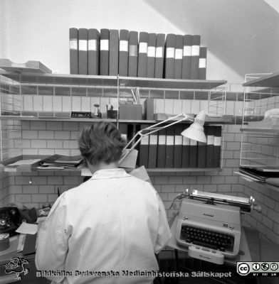 Lasarettet i Lund år 1967. Öronklinikens expedition.
Lasarettsfotograferna. Lund. Pärm "Div. tagningar, 1960 och t.v.". Sekreterare 1967  i ett trångt kakelklätt utrymme med snedtak på översta våningen på öronkliniken; rimligen urssprungligen ett hygienutrymme. Från negativ.
Nyckelord: Lund;Lasarett;Universitet;Universitetssjukhus;USiL;Öron;ÖNH;Öron-näsa-hals;Otologi;Otiatri;Administration;Sekreterare;Expedition