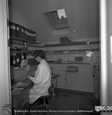 Lasarettet i Lund år 1967. Öronklinikens expedition.
Lasarettsfotograferna. Lund. Pärm "Div. tagningar, 1960 och t.v.". Sekreterare 1967  i ett trångt kakelklätt utrymme med snedtak på översta våningen på öronkliniken; rimligen urssprungligen ett hygienutrymme. Från negativ.
Nyckelord: Lund;Lasarett;Universitet;Universitetssjukhus;USiL;Öron;ÖNH;Öron-näsa-hals;Otologi;Otiatri;Administration;Sekreterare;Expedition