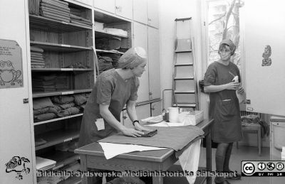 Interiörer från gamla kvinnokliniken i Lund, inför flytt till nya lokaler
Sjukhusfotograferna i Lund. Pärm Negativ, S/V. 1976.	38. Två sköteerskor eller biträden i textilförrådet på gamla kvinnoklinikens operationsavdelning. Från negativ. 
Nyckelord: Lasarett;Lund;Universitet;Universitetssjukhus;USiL;Kvinnoklinik;Gynekologi;Förråd;Textilier;Operationsavdelning