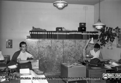 Två anonyma kontorister på Lasarettet i Lund 1960
Pärm Lasarettsfoto, interiörer och exteriörer m.m. 1958 - 1960. 17. Lokaler, Las. 1960. Kontorister på lasarettskontoret vid valven på Allhelgona Kyrkogata. Foto inför administrationens flytt till norra lasarettsområdet. Bild SMHS282 kommer från samma negativ, men har ingen ytterligare märkning.	Från negativ
Nyckelord: Lasarettet;Lund;Universitet;Universitetssjukhus;USiL;Administration;Kontor;Kontorist