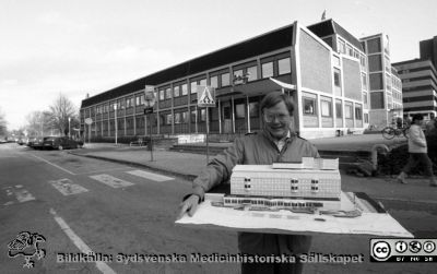 En arkitektmodell av tillbyggnaden av administrationshuset i bakgrunden, 1986
Sjukhusfotograferna i Lund. Pärm Negativ S/V, färg-1986. Chefen för näradministrationen vid Lasarettet i Lund, Leif Granath, med en arkitektmodell av den kommande utbyggnaden av administrationshuset. Film 109-1986. Foto: 86-11-07. Från negativ. 
Nyckelord: Lasarettet;Lund;Universitetssjukhus;USiL;Administration;Utveckling