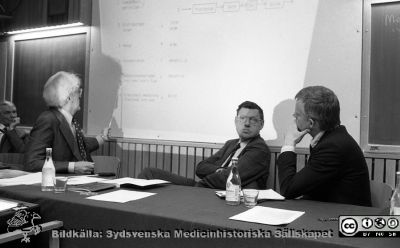 Disputation på kirurgiska kliniken i Lund 1987
Sjukhusfotograferna i Lund. Pärm Negativ, Dia, 1987. 70/1987. Professor Dag Hallberg, Huddinge som opponent längst till vänster och klinikchefen professor Stig Bengmark i bildens mitt. Disputanden Mogens Bugge ses til höger. Han flyttade sedan till Göteborg. Avhandlingen berörde serotoninomsättning i hjärnan vid experimentell modell med hepatectomi på råtta. På väggen visas ett schema över serotoninbildning från tryptophan. Från negativ. 
Nyckelord: Lasarettet;Lund;Universitetssjukhus;USiL;Kirurgisk;Klinik;Disputation;Avhandling;Utbildning