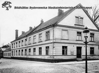 Den äldsta bevarade byggnad som tillhört Lasarettet i Lund
Kurhuset av 1824 i hörnet av Sandgatan och Paradisgatan, med tillbyggnaden från 1864 (cura posterior). Tvätt- och maskinhuset längre bort är från 1874, portvaktshuset från 1887. Bakom detta skymtar patologiska institutionen från 1887. På kurhusets gavel ses poliklinikskyltar. Tidigare fanns här också polikliniker för barn och invärtes- kvinnor- öron-näs och halssjukdomar. Gatlyktan har gasljus.
Lasarettsarkivet. Publicerad på sidan 110 i Albert Flaums historik 1968 över Lasarettet i Lund.
Nyckelord: Äldsta;Första;Byggnad;Kurhus;Lasarettet;Lund;Södra;Området;Portvakt;Portvaktshus;Patologisk;Institution;ÖNH;Barn;Poliklinik;Medicin;Gynekologi;