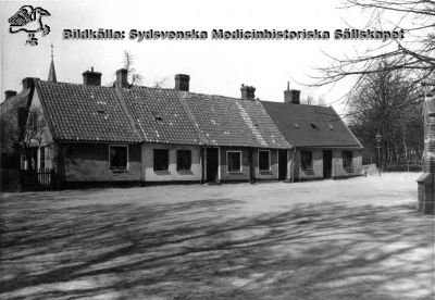 Gatubild från Lund nära 1900; Allhelgona Kyrkogatas östra ände vid biskopshuset
Kanske är det Allhelgonakyrkans torn som sticker upp i bakgrunden, och rimligen låg därmed husen i norra delen av nuvarande södra lasarettsområdet, ungefär där gamla entréhuset och östra valvet nu ligger. Husen revs när lasarettsområdet utvidgades norrut i början av 1900-talet. Den mur eller murade staketstope som skymtar i bildens högra kant kan höra till gamla biskopshuset. Solens skuggor antyder att längan låg i huvudsak i nord-sydlig riktning. 
Nyckelord: Kapsel 01;Regionarkivet;Lund;Lasarettet;Universitet;Allhelgona;Kyrkogata;Universitetssjukhuset;Söder