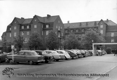 Ursprungliga ortopediska kliniken i Lund
Huset byggdes c:a 1930 och rymmer sedan 1970-talet kvinnokliniken. 
Bilarnas årsmodeller och den kvarstående utskjutande baldakinen på Jubileumskliniken / Onkologiska kliniken antyder att bilden tagits på tidigt 1960-tal. - Foto monterat på kartong.
Nyckelord: Kapsel 02;Regionarkivet;Lasarettet;Lund;Ortopedi;Ortopedisk;Klinik