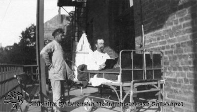 Patient på lungklinikens balkong 1918
Påskrift: "Tillhör Otto Roos, Kung Oscars väg 9a, Lund. Lungklinikens balkong omkr. 1918". Foto, monterat. Lungsjuka patienter behandlades vid denna tid med bra mat, vila, strikt sängläge, frisk luft och solljus.
Otto Roos var född 1904 och dog 1992, och  var vid sin död bosatt på adressen ovan. Rimligen är det inte han som syns på bilden, för både vårdaren och patienten är äldre än 14 år. Gissningsvis är det hans far eller möjligen en äldre bror som syns på bilden.
Nyckelord: Kapsel 02;Regionarkivet;Lasarettet;Lund;Lungklinik;Söder;Södra;Område