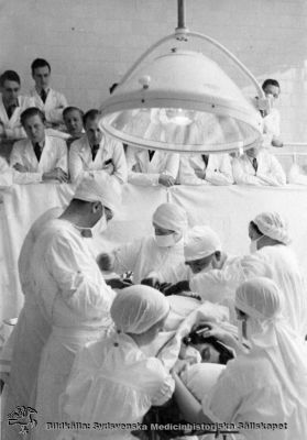 Kirurgiska klinikens operationsavdelning i mars 1937
Påskrift: Kir. op. Mars 1937. Åke Rydén, foto. Rimligen Gustaf Petrén som operatör (sittande). Omonterat foto.
Narkossystern droppar eter på öppen mask. Operationspersonalen är fullt sterilklädd med mössa och munskydd, men åskådarna (studenterna) behövde ännu bara bära vit rock. Färgade operationskläder infördes först ett par decennier senare.
Nyckelord: Kapsel 02;Regionarkivet;Lasarettet;Lund;Kirurgi;Kirurgisk;Klinik;Operation;Gustaf,Petrén;