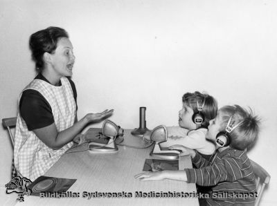 Undervisning av hörselskadade barn.
Foto monterat på kartong. Påskrift: "Personal LL. Hörselskadade på skolundervisning. Foto i Lasarettsarkviet". Apparaturen antyder mitten på 1900-talet.
Nyckelord: Kapsel 04;Foto;Monterat;Personal;Lund;Lasarett;Skola;Hörselskadade;Barn