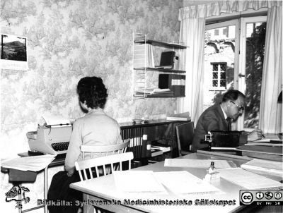 Lasarettsadministrationen i Lund 1960
Omonterat och omärkt foto, i mapp från sjukhusdirektörens kontor med påskrift "Fotoarkiv. Paradisgatan + annat gammalt". Foto troligast från sjukhuskontoret vid Valvet på södra lasarettsområdet. Kanske Per-Börje Jönsson vid skrivbordet. Bokhyllan på väggen är en s.k. String-hylla, populär på 1950-talet. Räknesnurran är handvevad, och skrivmaskinen är nog av mekanisk typ. Foto 1960, kanske inför avflytten till norra lasarettsområdet c:a 1960. 
Nyckelord: Lasarettet;Lund;Universitet;Universitetssjukhus;USiL;Administration;Kontor;Kanslist