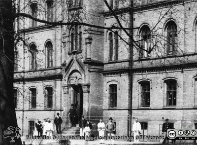 Ögonkliniken från år 1884. 
I ögonklinikens källarvåning fanns lasarettets första egna köksinrättning, våningen 1 tr upp kontorslokaler. Uppe på trappan står sysslomannen P. Lundborg. Foto 1903 av Lina Jonn. Foto: Lunds universitetsbibliotek. Originalfoto, monterat
Nyckelord: Kapsel 17;Lund;Lasarett;Södra;Område;Lasarettsområde