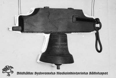 Vällingklocka från Lasarettet i Lund
Omärkt bild. Originalfoto. Monterat. S.k. vällingklocka. Påskrift på klockan: J.P.J Chanzon. Lunds Lazarett. MDCCCLXXX. (dvs 1880).
Klockan användes från början på 1800-talet vid Lunds lasarett för matsignal av spisvärden, en syssla som upphörde 1882 då lasarettet fick ett eget kök. Klockan hängde bl.a. i en alm nära Paradisgatan på södra lasarettsområdet, och ännu 1968 kunde man se märken efter den i trädet (Flaum 1968, sid 122-123). Den finns nu på Kulturen i Lund och kallas där "matklocka" (KM 81820).
Nyckelord: Kapsel 18, Inventarier