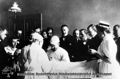 Prof. J. Borelius opererar omkring år 1900
Den första operationssköterskan, Sophiasystern Hedvig Olander, assisterar. Märk gaslampan. Narkosutrustningen består av eterflaska, bomullstuss och en mask. Aseptiken har slagit igenom så till vida att operationspersonalen bär skyddskläder. De medicine kandidater är emellertid iförd mörk kostym. Syster Olander bär mössa men saknar handskar. Foto erhållet av prof. Borelius dotter, fru Klara Welin. Publicerad i Flaum 1968, sid.189. Originalfoto Monterat
Nyckelord: Kapsel 18;Interiör;Operation;Lund;Lasarett;Kirurgi