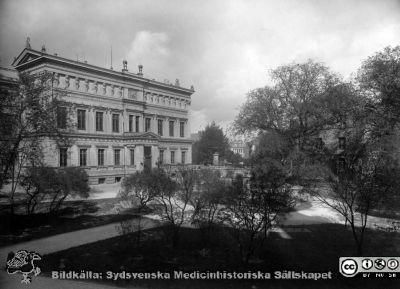 Utsikt mot Lundagård med universitetsbyggnadens söderfasad och Kungshusets västerfasad bakom träden. 
Ur ett fotoalbum som visar den lägenhet på Kyrkogatan 15 där Gustaf Ahlström bodde under sin tid som professor i oftalmiatrik i Lund (1911 - 1927). Bilderna bör rimligen vara tagna under 1920-talet, någon tid innan han vid sin pensionering 1927 flyttade till Göteborg. 
Nyckelord: Gustaf;Ahlström;Album;Professor;Oftalmiatrik;Lund;Kyrkogatan;Lägenhet