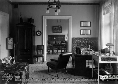 Troligen mottagningsrum i Gustaf Ahlströms lägenhet i Lund under hans tid som professor i oftalmiatrik i Lund 1911-1927 
Synprövningstavla till vänster i fonden, med avskärmad belysning på stativ framför. Instrument, lösningar. läkemedel och mottagningsutrustning i monter till höger. Ett par ögoninstrument på stativ på bordet i bortre rummet, rimligen använt som mörkrum. Instrumentet till höger är en tidig version av Gullstrands spaltlampa, introducerad c:a 1911. Ur ett album med bilder från Gustaf Ahlströms hem, rimligen från 1920-talet.
Nyckelord: Album;Gustaf;Ahlström;Professor;Oftalmiatrik;Lund;Kyrkogatan;Lägenhet