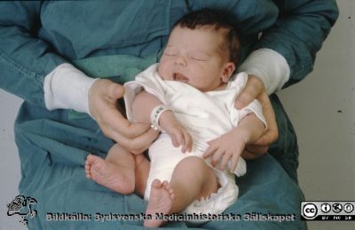 Nyfött barn i op-klädd persons knä på kvinnoklinikens förlossningsavdelning 1984
Ur låda med blandade diabilder från sjukhusfotograferna i Lund, 1984. 
Nyckelord: Lund;Lasarett;Universitet;Universitetssjukhus;KK;Kvinnoklinik;BB;Förlossningsavdelning