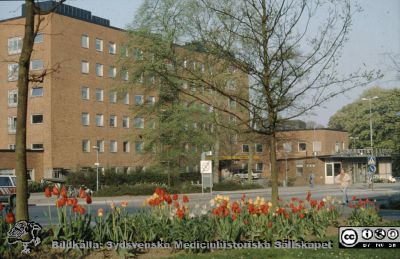 Barnkliniken vid huvudentrén med portvaktshuset, vid Getingevägen 1989.
Ur låda med blandade diabilder från sjukhusfotograferna i Lund, 1970-, 1980- och 1990-talen. Lasarettet i Lund 1989. 
Nyckelord: Lasarettet;Lund;Universitetssjukhuset;USiL;Barnklinik;Pediatrisk;Klinik;Portvakt;Entr