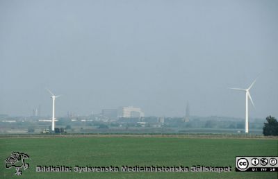 Vårligt soldis över lundaslätten vid Lunds Energis (nu Kraftringens) vindkraftverk vid Värpinge. 
Lasarettet i Lund i bakgrunden. Helikopterplattan på taket är ännu inte byggd. Kylvattentornet vid Lunds Energis (nu Kraftringens) kraftvärmeverk har byggts. Foto troligast på 1990-talet.
Nyckelord: Lasarett; Lund; Universitet; Universitetssjukhus; Exteriör;Lundaslätten