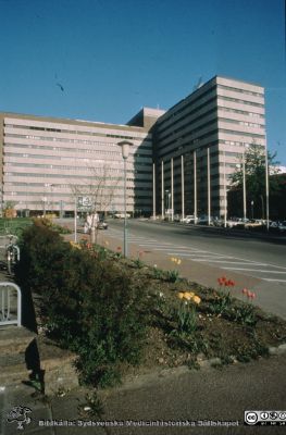 Infarten mot centralblocket på Lasarettet i Lund en tidig vårdag före 1986
Ur låda med blandade diabilder från sjukhusfotograferna i Lund, 1970-, 1980- och 1990-talen.  Det glasade entrépartiet (byggt 1986) på husets västerfasad är ännu inte på plats och satellitantennerna på husets tak har inte satts upp.
Nyckelord: Lund;Lasarett;Universitetssjukhus;Fasad;Entr;Infart;Gata