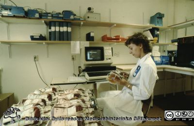 Interiör på blodcentralen i Lund
Ur låda med blandade diabilder från sjukhusfotograferna i Lund, 1970-, 1980- och 1990-talen. Bild utan påskrift. Blodpåsar kontrolleras. Rimligen på blodcentralen.

