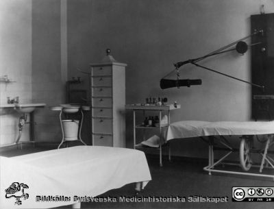 Operations- eller behandlingsrum på ögonkliniken i Lund i mitten på 1920-talet. 
Till höger invid väggen ett höj- och sänkbart operationsbord. Till vänster ett ställ med en spann och handfat tvagnings- och sköljfat av den typ som användes vid denna tid, liksom tvättställ på väggen med rinnande vatten.
Nyckelord: Ögon;Klinik;Lasarettet i Lund;Medicinska fakulteten;Operationsrum;Behandlingsrum