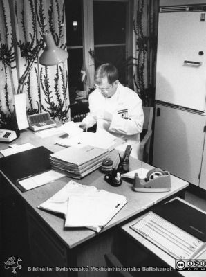 Docent Magnas Hägerdal i arbete på Lasarettet i Lund på 1980-talet
Överläkare docent Magnus Hägerdal i arbete med pappersjournaler på Lasarettet i Lund, rimligen på 1980-talet. Mörkret har fallit på utanför. Telefonen är av bordsmodell, liksom den mer fyrkantiga och högtalande s.k. snabbtelefonen. Någon dator är inte i sikte. På skrivbordet står en liten flaska vit färg ("Tipp--Ex") som användes för att måla över text vid rättning i skrivelser och motsvarande. I journaler fick sådant inte användas, det var bara överstrykningar som dög där, så att den gamla texten fortfarande gick att läsa om så behövdes. Foto Krister Malmström, tidningen Arbetet.
Nyckelord: Lasarettet;Lund;Universitetssjukhuset;USiL;Anestesiologiska;Narkos;Kliniken;Avdelningen;Intensivvård