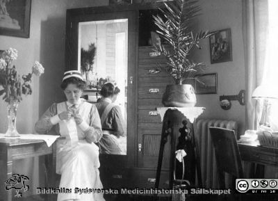 En ung sjuksköterska med SSSH-mössa och brosch sitter med handarbete i sitt rum.
Bild 28 i husmor Anna Landelius (1880-1964) efterlämnade fotoalbum. Ingrid Frennhoff (f. 1944) hade övertagit detta album efter sin morfar som i sin tur tagit hand om det efter sin syster husmor Anna på Lasarettet i Lund. Gåva till Sydsvenska Medicinhistoriska Sällskapet nyåret 2020 - Rimligen en vän till Anna Landelius. Foto kanske på 1920-talet.
Nyckelord: SSSH;Södra Sveriges Sjuksköterskehem;Sjuksköterska