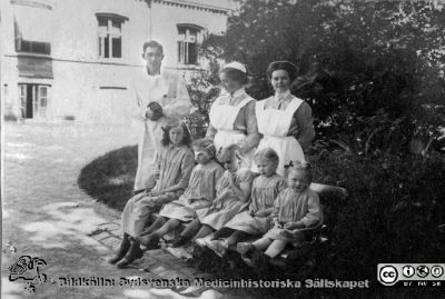 Barnaskara i en park på Lunds lasarett i kv. Paradis i Lund. 
Bild 048-1 i husmor Anna Landelius (1880-1964) efterlämnade fotoalbum. Ingrid Frennhoff (f. 1944) hade övertagit detta album efter sin morfar som i sin tur tagit hand om det efter sin syster husmor Anna på Lasarettet i Lund. Detta album är en gåva från Ingrid Frennhoff till Sydsvenska Medicinhistoriska Sällskapet nyåret 2020.

Kanske ögonpatienter. I bilden ses en undersköterska med ett St Georgskors som brosch i kragen, en sköterskeelev med s.k. elevsnibb på huvudet, möjligen Anna Landelius, samt en icke identifierad läkare.
Nyckelord: Patienter;Barn;Sköterskor;Läkare