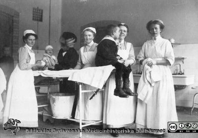 Vårdpersonal och patienter i en polikliniksal, troligen på kirurgen i Lund. 
Bild 53 i husmor Anna Landelius (1880-1964) efterlämnade fotoalbum. Ingrid Frennhoff (f. 1944) hade övertagit detta album efter sin morfar som i sin tur tagit hand om det efter sin syster husmor Anna på Lasarettet i Lund. Detta album är en gåva från Ingrid Frennhoff till Sydsvenska Medicinhistoriska Sällskapet nyåret 2020.

Sköterskan längst till höger bär SSSH-mössa och brosch.Troligen står Anna Landelius mitt i bilden, men bär ännu inte SSSH-mössa och brosch, d v s hon är fortfarande under utbildning. Hon utexaminerades 1914-04-01. Foto alltså troligen på 1910-talet.
Nyckelord: Sköterskor;Sjuksköterskor;Patienter;Barn;Poliklinik