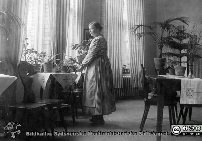 Kvinnlig patient med vattenkanna i dagrum
Bild 059-1 i husmor Anna Landelius (1880-1964) efterlämnade fotoalbum. Ingrid Frennhoff (f. 1944) hade övertagit detta album efter sin morfar som i sin tur tagit hand om det efter sin syster husmor Anna på Lasarettet i Lund. Detta album är en gåva från Ingrid Frennhoff till Sydsvenska Medicinhistoriska Sällskapet nyåret 2020.

Troligen ett dagrum på kirurgiska kliniken på Lasarettet i Lund i kv. Paradis. Värmeelementen i förnsersmygarna anger att fotot togs under första halvan av 1900-talet efter införandet 1905 av vattenburen centralvärme.
Nyckelord: Patient;Kvinna;Dagrum;Lasarettet i Lund;Paradis;vattenkanna