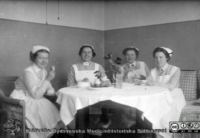 Fyra sköterskor på kaffepaus. 
Bild 067-5 i husmor Anna Landelius (1880-1964) efterlämnade fotoalbum. Ingrid Frennhoff (f. 1944) hade övertagit detta album efter sin morfar som i sin tur tagit hand om det efter sin syster husmor Anna på Lasarettet i Lund. Detta album är en gåva från Ingrid Frennhoff Sydsvenska Medicinhistoriska Sällskapet nyåret 2020.
En av sköterskorna bär armbindel med kors på. En har en rund brosch i kragen. Ingen är SSSH-sköterska. Rimligen ingår de i Anna Landelius bekantskapskrets.
Nyckelord: Sjuksköterskor;Kaffepaus