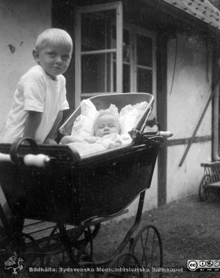 Liten pojke vid barnvagn med spädbarn i. 
Bild 069-2 i husmor Anna Landelius (1880-1964) efterlämnade fotoalbum. Ingrid Frennhoff (f. 1944) hade övertagit detta album efter sin morfar som i sin tur tagit hand om det efter sin syster husmor Anna på Lasarettet i Lund. Detta album är en gåva från Ingrid Frennhoff till Sydsvenska Medicinhistoriska Sällskapet nyåret 2020.
Kanske syskonbarn till Anna Landelius på hennes föräldragård Jakobslund i Ljungsbro.
Nyckelord: Syskonbarn;Föräldragård;Spädbarn;Barnvagn