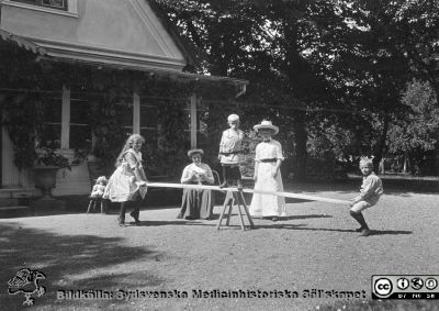 Barn leker med en gungbräda på gårdsplan på lantgård. Två kvinnor är med dem. 
Bild 73 i husmor Anna Landelius (1880-1964) efterlämnade fotoalbum. Ingrid Frennhoff (f. 1944) hade övertagit detta efter sin morfar som var bror till husmor Anna på Lasarettet i Lund. Detta Album har skänkts till Sydsvenska Medicinhistoriska Sällskapet.

Kläderna talar för att fotot togs en solig och varm dag under 1900-talets första decennium. Kanske på Anna Landelius fädernegård Jakobslund i Ljungsbro?
Nyckelord: Barn;Lantgård;Gungbräda