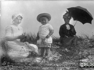 Strandbild med kvinnor och barn. Anhöriga till Anna Landelius.
Bild 84 i husmor Anna Landelius (1880-1964) efterlämnade fotoalbum. Ingrid Frennhoff (f. 1944) hade övertagit detta album efter sin morfar som i sin tur tagit hand om det efter sin syster husmor Anna på Lasarettet i Lund. Detta album är en gåva från Ingrid Frennhoff till Sydsvenska Medicinhistoriska Sällskapet nyåret 2020.
Den äldre kvinnan bär mörka kläder, utstyrd hatt och parasoll. Den ljusklädda yngre kvinnan har en kikare i händerna. Foto på en badstrand, troligen i Skåne och nära sekelskiftet 1900.
Nyckelord: Strandbild;Barn;Parasoll;Kikare