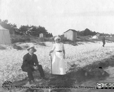 Man och kvinna i sommarkläder vid badstrand med strandtält och badhytter. Rimligen anhöriga till Anna Landelius.
Bild 87 i husmor Anna Landelius (1880-1964) efterlämnade fotoalbum. Ingrid Frennhoff (f. 1944) hade övertagit detta album efter sin morfar som i sin tur tagit hand om det efter sin syster husmor Anna på Lasarettet i Lund. Detta album är en gåva från Ingrid Frennhoff till Sydsvenska Medicinhistoriska Sällskapet nyåret 2020.
 Falsterbo?
Nyckelord: Badstrand;Badhytter;Strandtält