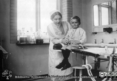 Sköterskeelev med liten pojke på brits på undersökningsrum, möjligen på kirurgiska kliniken i Lund.
Bild 26 i husmor Anna Landelius (1880-1964) efterlämnade fotoalbum. Ingrid Frennhoff (f. 1944) hade övertagit detta album efter sin morfar som i sin tur tagit hand om det efter sin syster husmor Anna på Lasarettet i Lund. Gåva till Sydsvenska Medicinhistoriska Sällskapet nyåret 2020.

Nyckelord: Sjuksköterskeelev;Barn;Undersökningsbrits;Kirurgiska kliniken