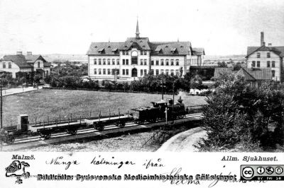 Vykort O. E. K. 8026 från Saumuel Bengtssons missionsbokhandel med bild av Malmö Allmänna Sjukhus nära sekelskiftet 1900.
 Ett godståg går på Malmö-Ystads Järnvägs spår alldeles hitom sjukhuset. Kortet är adresserat till Fröken Elisabeth Gyllenberg, Allmänna Sjukhuset, Södervärn (rimligen en dotter till sjukhusets syssloman E. A. Gyllenberg). Vykortets poststämpel är inte tolkningsbar, men den felaktiga postadressen (Södervärn i stället för Malmö och ingen gatuadress) pekar på att kortet skickats inom Malmö eller i varje fall Skåne.
Foto från en samling funnen på KK i Lund hösten 2016 och tillvaratagen av en sekreteraren där (Elisabet Gyllander) i samband med att hon gick i pension. Hon överlämnade bilderna till Sydsvenska Medicinhistoriska Sällskapet där hon tyckte de passade bättre än hos henne.  
Nyckelord: Malmö;Allmänna;Sjukhus;MAS;Järnväg;Fasad;Exteriör;Vykort
