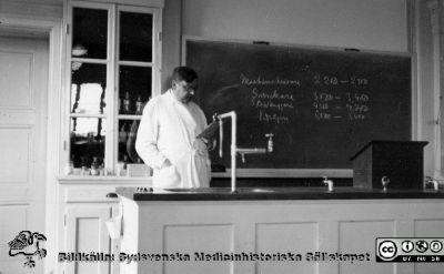 Erik M. P. Widmark, professor i medicinsk och fysiologisk kemi från 1920
Erik M. P. Widmark i en föreläsningssal på 1924 års institution för medicinsk kemi i Lund. På tavlan ett antal yrkestitlar (Maskinskrivare, Snickare, ...huggare, Vedsågare) och kanske en kostnadsklkyl för respektive yrke. Föreläsningssalen liknar den i bild 180615-07. Bildkälla barnbarnet Eva Marie Widmark. Foto privat, tidigast 1924  Fotograf okänd.
Nyckelord: Medicinsk och fysiologisk kemi;Medicinska fakulteten;Lunds universitet;Förläsningssal