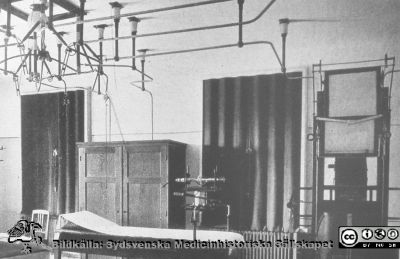 Röntgenrummet på Ängelholms sjukhus nära sekelskiftet 1900.
Efter diabild i sjuksköterskan Lillie Börjessons  samling från Ängelholms sjukhus. Sjukhuset fick röntgenutrustning redan 1907, och bilden visar en mycket tidig röntgenanläggning. Man ser röntgenröret ovanför britsens fotända och de oisolerade koppartrådarna som ledde den mycket högspända elektriciteten till det. Koppartrådarna sitter fast i taket med porslinsisolatorer. Den mycket högspända strömmen genererades vid denna tid av en induktionsspole som tjöt starkt och som därför ofta ställdes i ett skåp som det som syns i bilden.
Till höger i bild ses ett s.k. Forssellstativ för undersökning av patient i stående.
Nyckelord: Ängelholm;Engelholm;Sjukhus;Röntgen