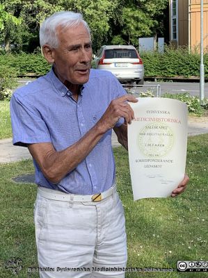 Lars Malm, nyutnämnd hedersledamot vid Sydsvenska Medicinhistoriska Sällskapets årsmöte den 10 juni 2021. 
Lars Malm har just utnämnts till hedersledamot i Sydsvenska Medicinhistoriska Sällskapets årsmöte den 10 juni 2021. Det avhölls i parken utanför Livets Museum en strålande vacker försommarkväll. Här visar Lasse stolt upp sitt diplom. Det står att han blivit korresponderande ledamot på det, men det var hedersledamot han blev. Lasse fick senare ett rättat diplom.
Nyckelord: Sydsvenska Medicinhistoriska Sällskapet;SMHS;Hedersmedlem;Fest;Pandemitid;rsmöte