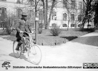 Verkmästare Johannes Hultén utanför södra fasaden till kirurgiska klinikens annex
Verkmästare Johannes Hultén på cykel utanför södra fasaden till kirurgiska klinikens annex. I cykelns ram sitter något som kanske är en motor, och cykeln är kanske en mycket tidig variant av vad som i modernare tid kom att kallas en moped? Kjell Edvardsson i Lund har hittat att Maskinfabriken Scania i Malmö (senare sammanslagen med Vabis i Södertälje) åtminstone 1903 tillverkade cyklar med en liknande hjälpmotor av franskt fabrikat (Clément), varav en finns bevarad på Scaniamuseet i Södertälje.
Verkmästare Hultén var intresserad av motorcyklar och ägde och körde senare större sådana.
Ur ett fotoalbum gjort av Johannes Hultén (1873-1951), verkmästare (elektricitet) på Lasarettet i Lund. Bildkälla dotterdottern Viveka Rasmusson 2018.
Nyckelord: Lasarett;Lund;Universitetssjukhus;Motorcykel;Kirurgisk;Klinik;Annex;Södra lasarettsområdet